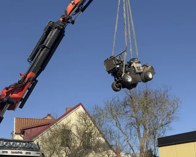 Kranbil lyfter en stubbfräs över en mur in till en trädgård